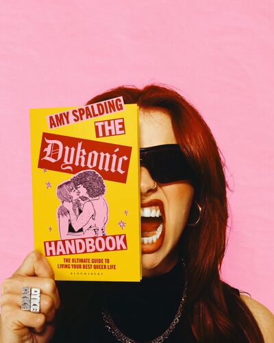 Amy Spalding holding up The Dykonic Handbook to her face - she wearing sunglasses, a black top and has her mouth open