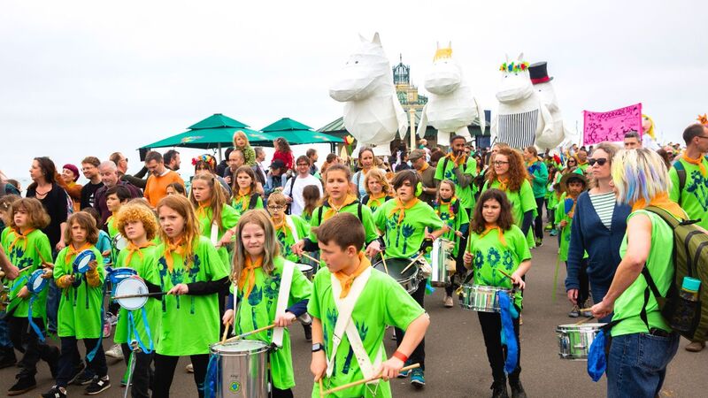 A school group of children in green t-shirts play instruments as they walk along Brighton seafront