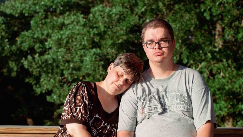 Two people are sat on a bench with trees and bushed behind them. The woman rests her head on the mans shoulder