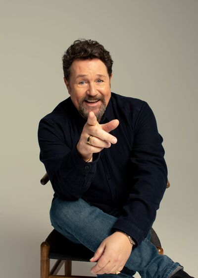 Michael Ball sitting on a chair with his finger pointing to the camera. He is smiling