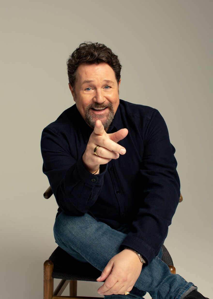 Michael Ball sitting on a chair with his finger pointing to the camera. He is smiling