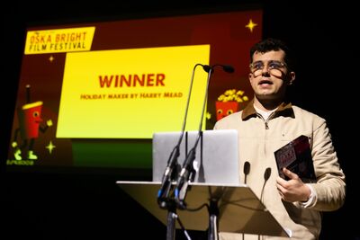 A young man stands behind a lectern holding an award. In the background a screen announces him as the winner 
