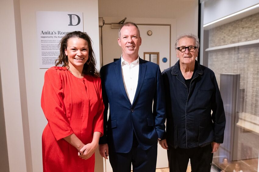 A white woman in a red dress, a white man in a blue suit and an older white man in a black outfit stand in front of the door of Anita's Room
