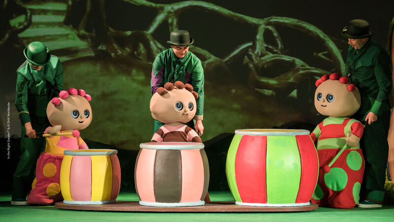 Three puppeteers dressed in green each control a round costumed multi-coloured figure, making them bang on stripey drums