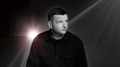 Kevin Bridges wearing a black shirt, with a spotlight behind him The image is black and white
