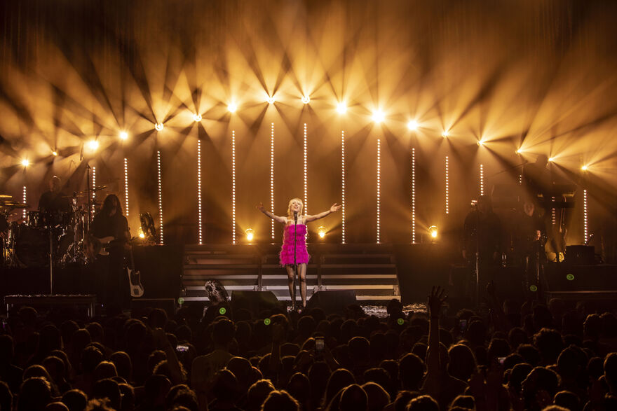 A blonde woman in a short, pink dress, stands on a stage with her arms held outwards. There is an arch of lights behind her. We can see the backs of peoples heads watching her.