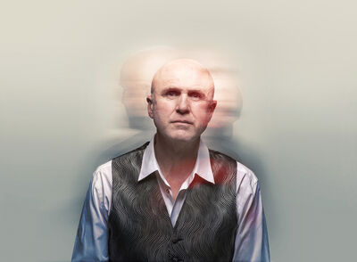A man in a grey waistcoat and white shirt. There are blurry photos of him behind.