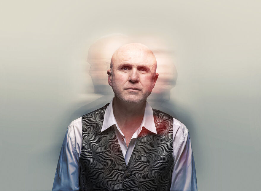 A man in a grey waistcoat and white shirt. There are blurry photos of him behind.
