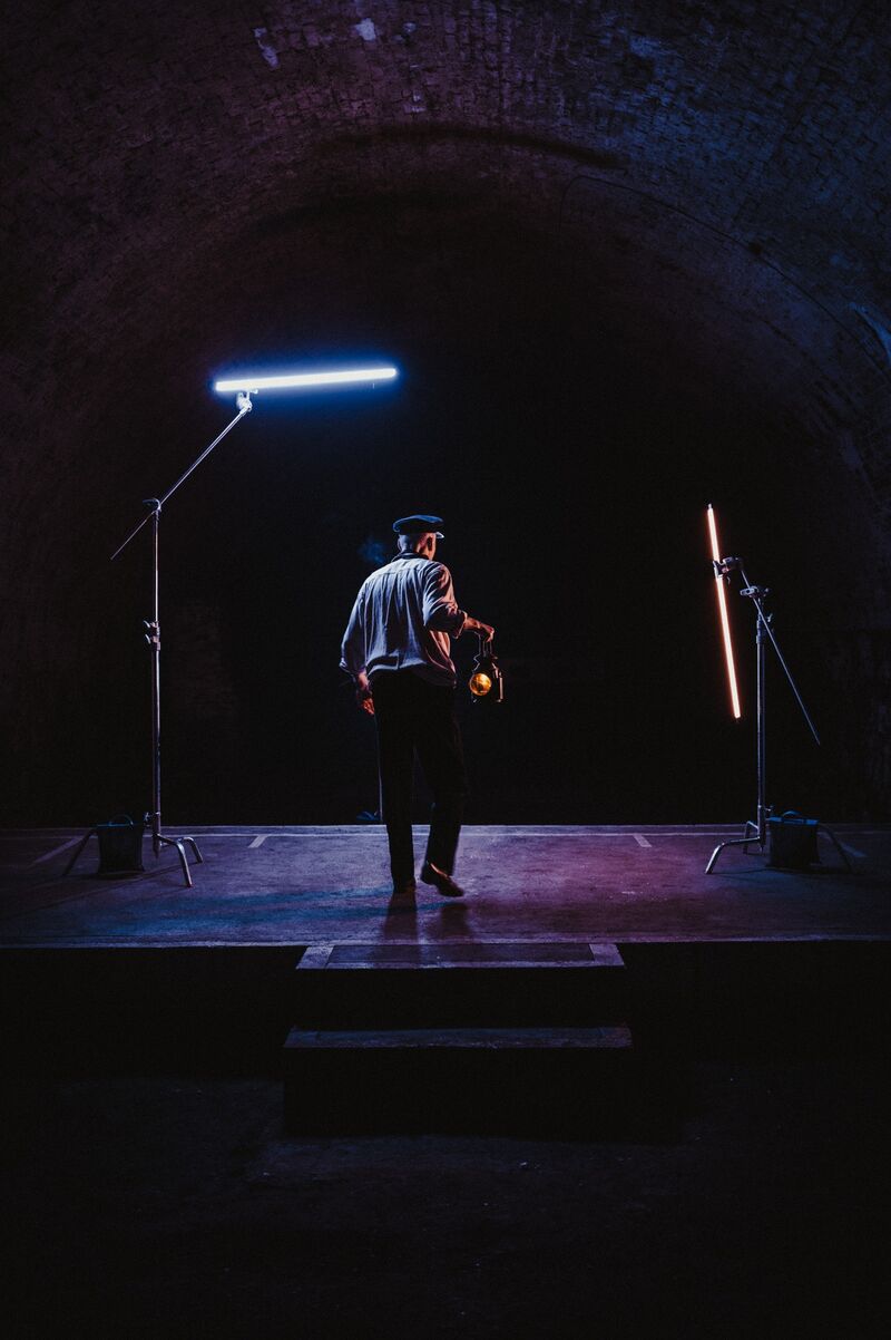 A man in a white shirt and a hat walks through a tunnel lit with neon lihgts. He holds a round orange lamp in his hand.