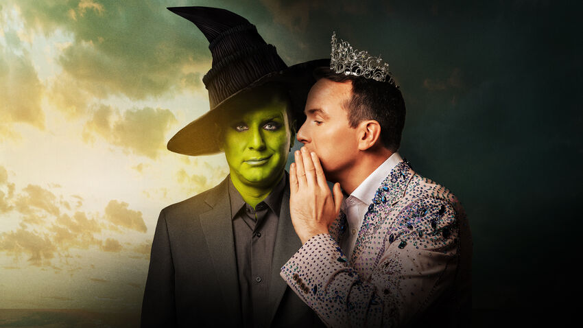 Simulating the Wicked cover, a man painted green faces the camera with a witches hat and a black suit. A man in a tiara and a sparkly suit is whispering in his ear. They are standing in front of a dark and cloudy background.