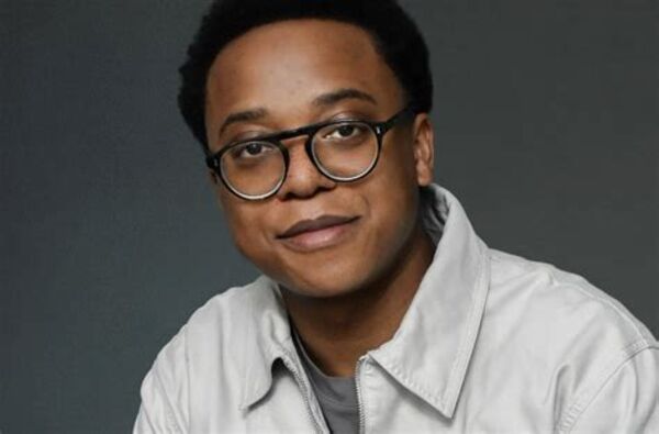 A black man with short black hair wearing dark rimmed glasses and a white zip-up jacket