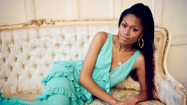 Isata Kanneh-Mason wearing a long blue dress and gold hoop earings, reclining on a chaise longue 