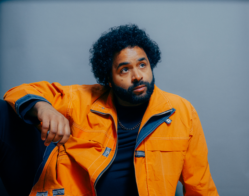 Steven Bamidele sat leaning against a wall looking thoughtfully in to the distance. He's wearing a casual orange jacket