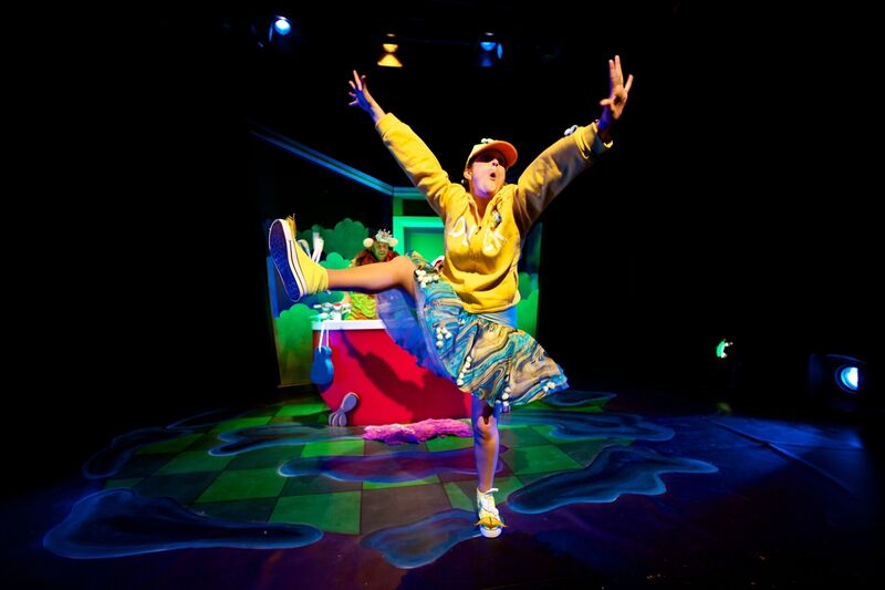 A woman wearing a bright yellow jumper and tutu stands on one leg with her arms in the air