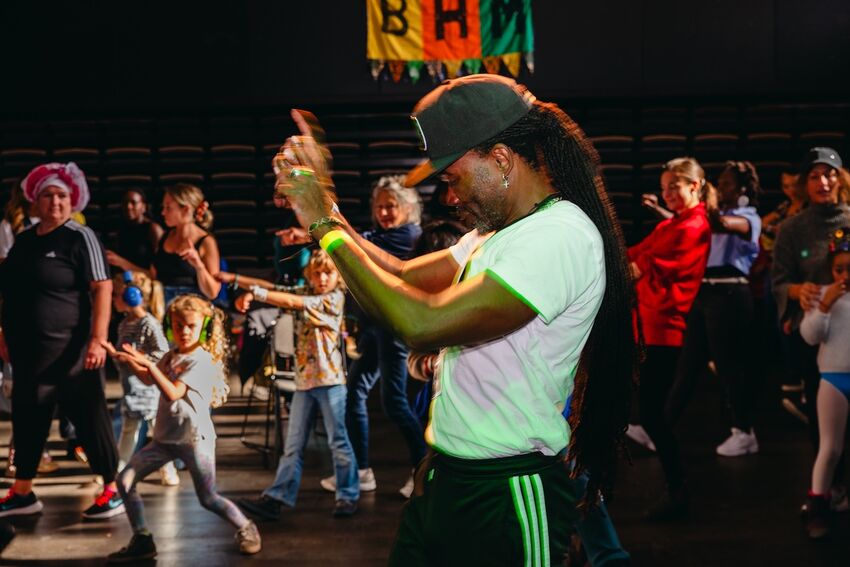 A man stands in front of a group of people dancing. He wears a white t-shirt, a black cap and had dreadlocks