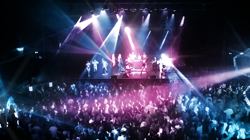 Large crowd standing in front of a stage with pink and blue lights around the venue