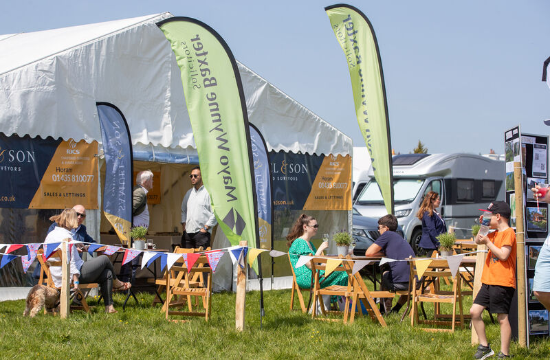 A white tent set up in a field surrounded by wooden folding chairs and mayo wynne baxter feather flags