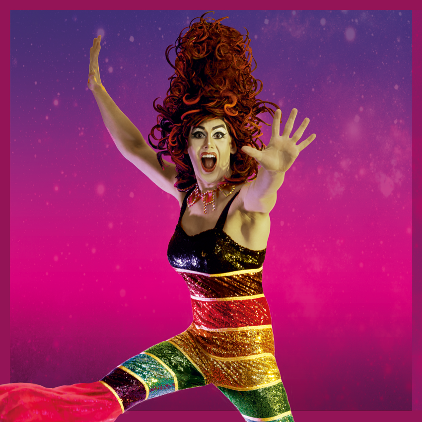 Aida H Dee jumping with arms oustretched. They're wearing a sparkly rainbow jumpsuit and have ginger curly hair in a beehive