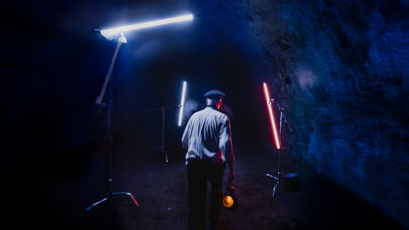 A man in a white shirt and a hat walks through a tunnel lit with neon lihgts. He holds a round orange lamp in his hand.