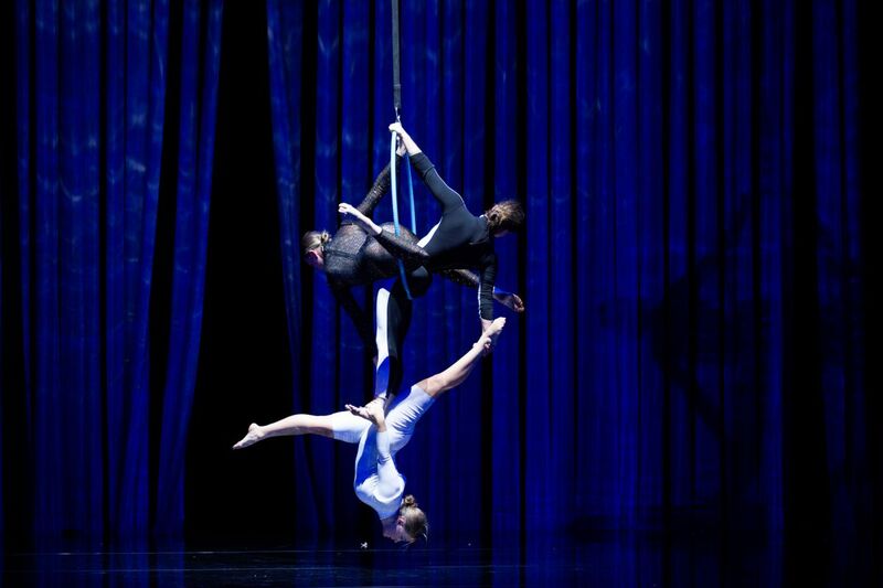 Three people are hanging on a rope. They are suspended in different positions, one person is in the splits upside down. They are in front of a dark blue curtain