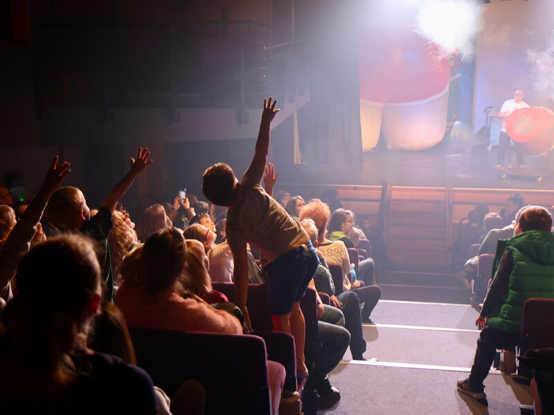 Children in the audience raise their hands with enthusiasm as they look towards a man on stage with a cannon