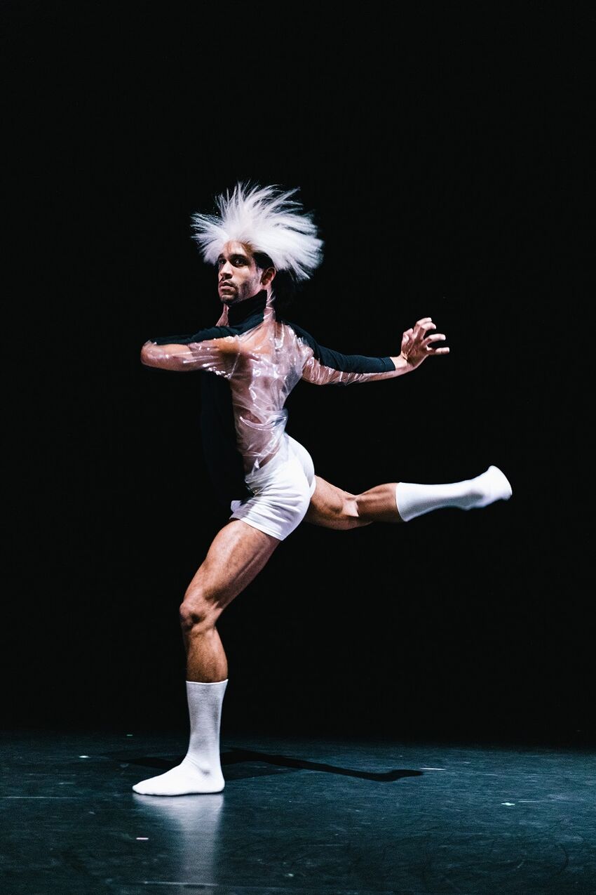 A man in white socks, white shorts and a sheer top stands on one leg, his other leg is raised and his arms our out. It appears he is mid pirouette. He wears a white wig on his head that is blowing as he twirls