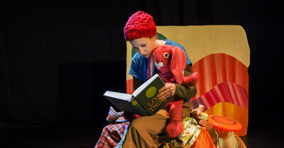 A person holding a stuffed red rabbit, reading it a book on stage. They are wearing a blue shirt and red hat