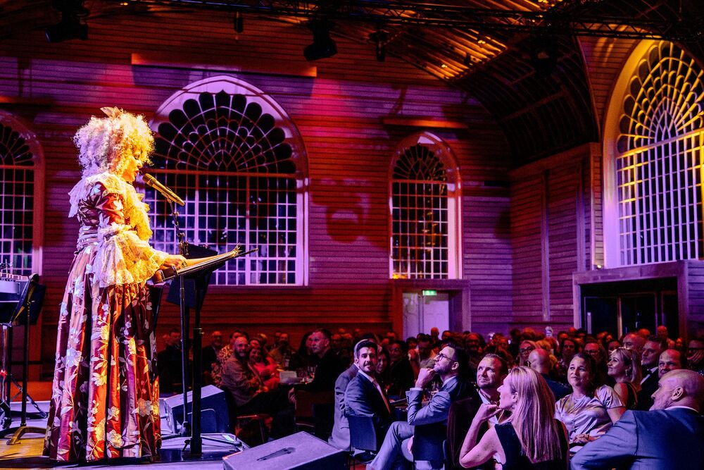 Cornucopia 2023. Maria Fitzherbert stands on stage talking to a crowd of diners in the Corn Exchange