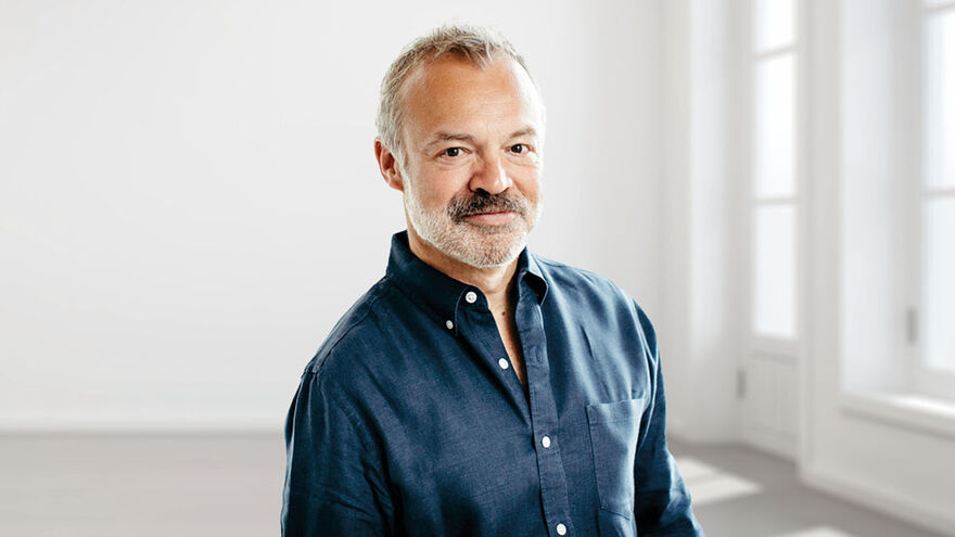 Graham Norton standing in a white room with large bright windows, wearing a dark blue shirt and smiling