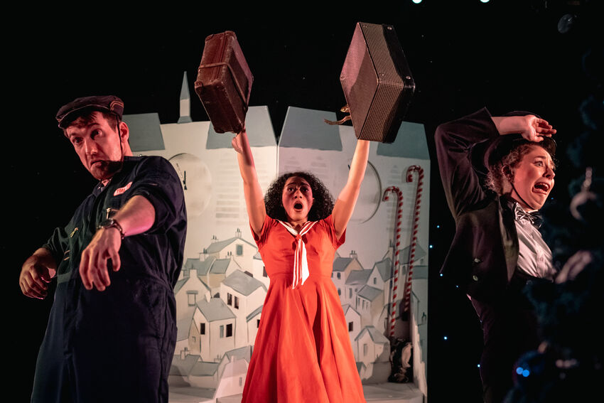 Three people on stage. There is a man wearing an all black outfit looking sad, a woman wearing a red 50s style dress and holding two brown bags in her hands, and there is a woman wearing a blazer and looking upset and confused