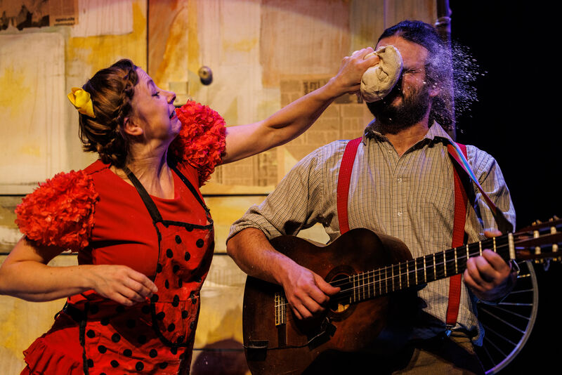 A woman dressed as a red hen throws bread dough into the face of a man playing the guitar