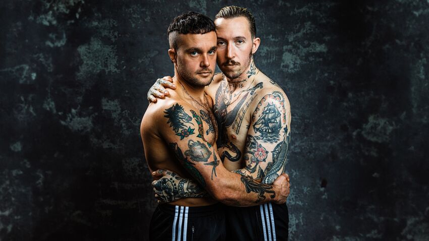 Two men embracing. Both are topless with tattoos and wearing tracksuit trousers