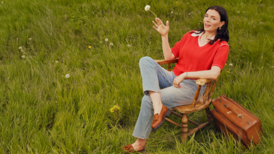 Aisling Bea sitting on a wooden chair in a field with a dandelion in her hand and her legs crossed