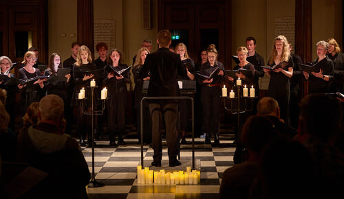 A choir of singers dressed in black sing facing their conductor with candles lit in front of them