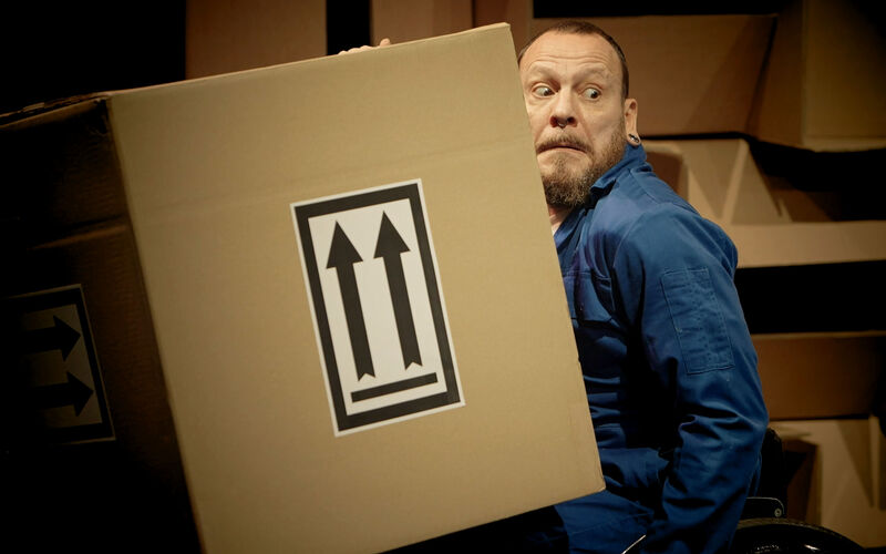 A man in a blue jumpsuit holds a big cardboard box with two upwards arrows on it. His face holds a worried expression.