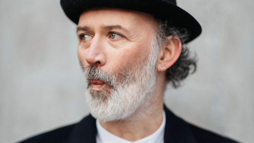 A white man with a grey beard stands against a grey wall. He is wearing a black bowler hat and a black jacket with a white shirt underneath