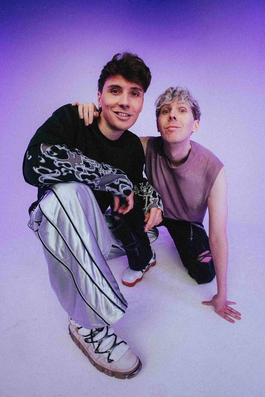 Two people pose together in a studio with soft purple lighting. One person crouches in front wearing a black top with patterned sleeves, shiny silver pants, and white sneakers, while the other kneels beside them in a sleeveless mauve shirt and ripped black jeans. They smile toward the camera against a plain light-colored backdrop.