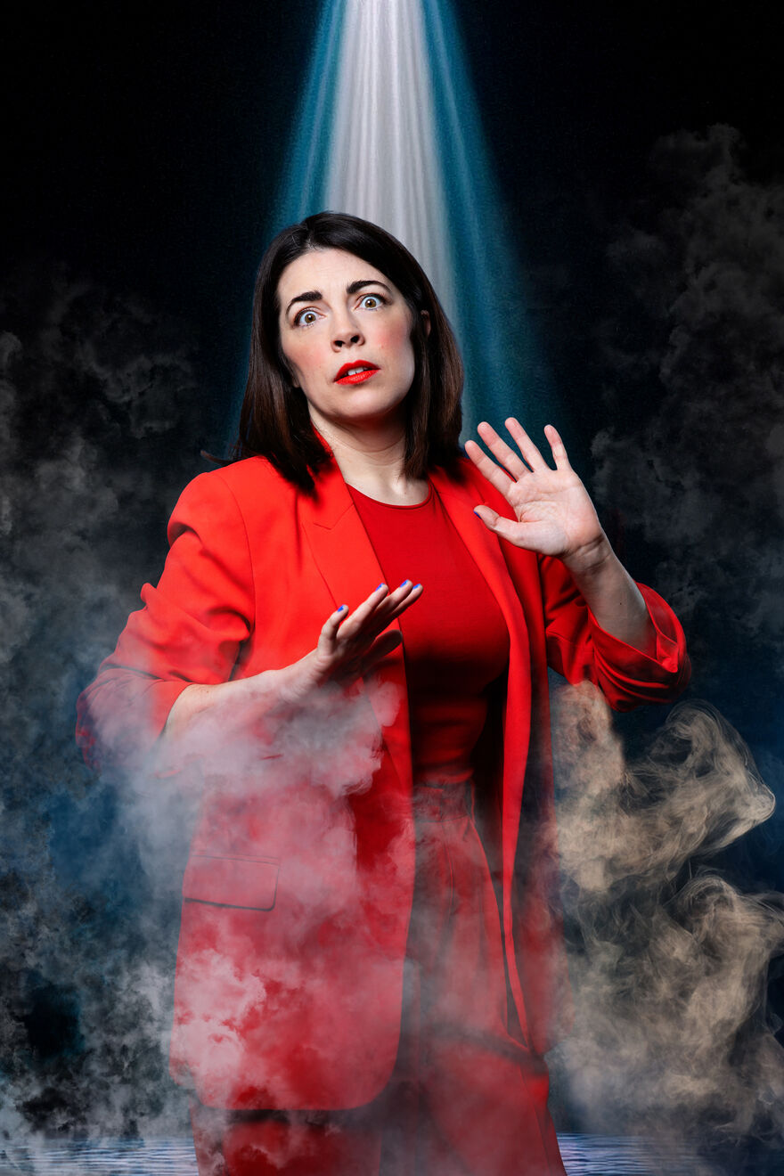 Rosie Holt is wearing an all red suit and she has a shocked expression. A mist surrounds her and a light is shining down on her