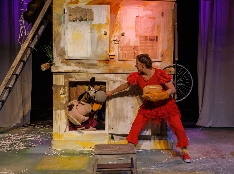 A woman dressed as a red hen reaches towards farmyard puppets to feed them bread from a loaf she's holding