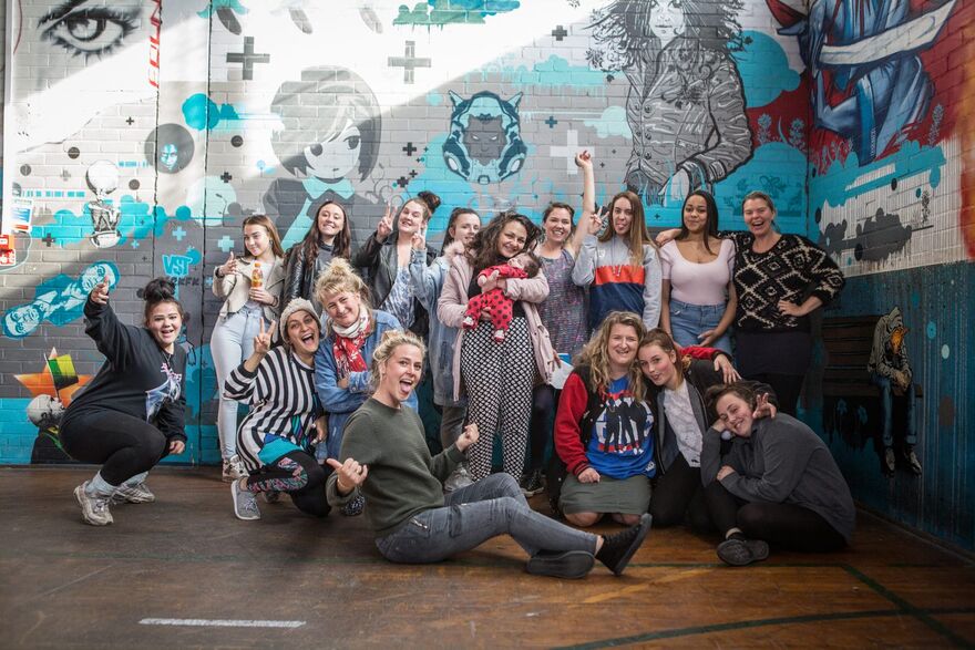 A group of 16 young women wearing casual clothing are posing in a group against a graffitied wall