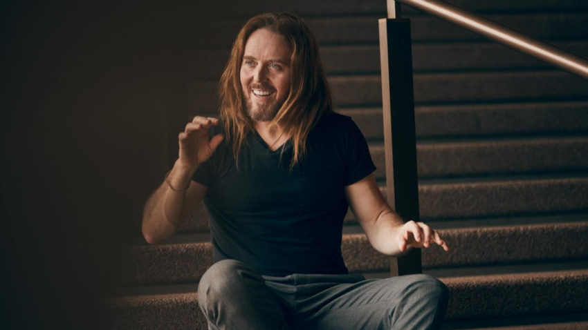 Tim Minchin sits laughing on a stairway with his hands outstretched animatedly