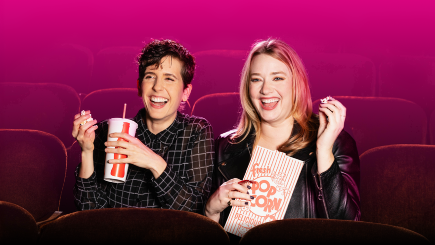 Two white women are sat in cinema chairs. One is holding a large drink and the other is holding a box of pop corn. Both women are laughing.