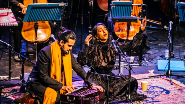 Two performers sit cross legged on stage in front of an orchestra. One is playing a traditional Indian instrument, the other is singing with her arms outstretched