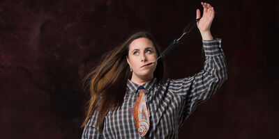 Kate Dolan wearing a plaid shirt and a trout tie. She has a fish hook in her mouth pulling upwards and she is looking to the left