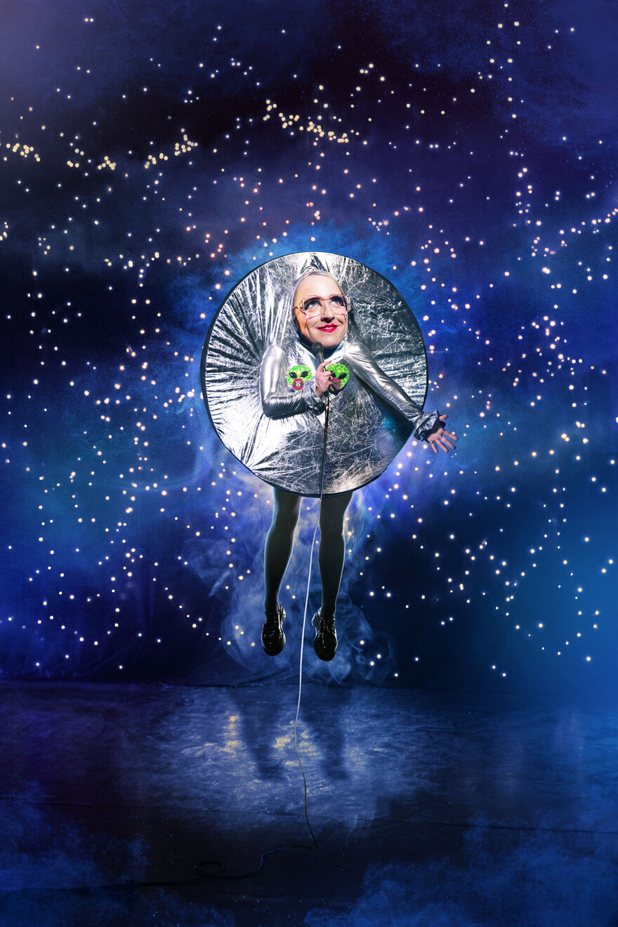 Lucy Pearman wearing a silver circle shape, holding two green alien heads, with a galaxy and star background.