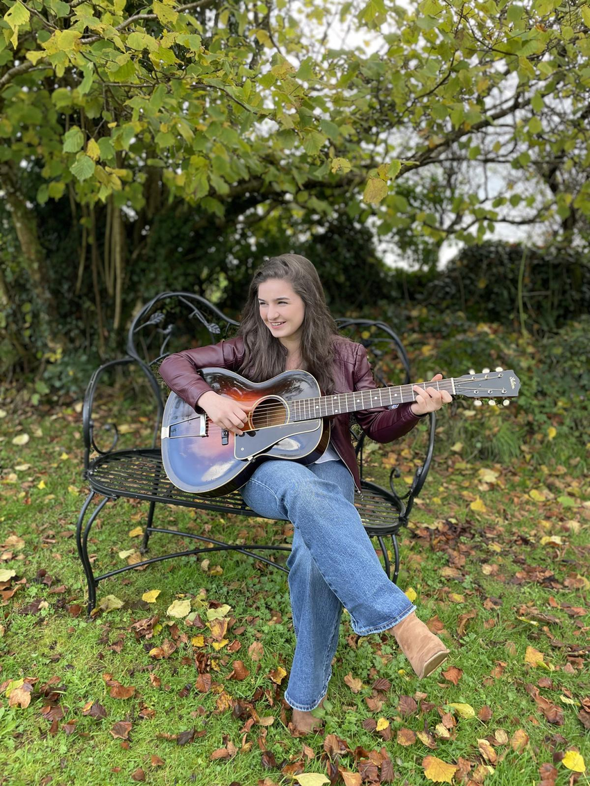 Muireann Bradley smiling as she sits on a park bench playing an acoustic guitar. She's a 17 year old with white skin and long dark hair, wearing jeans and a leather jacket
