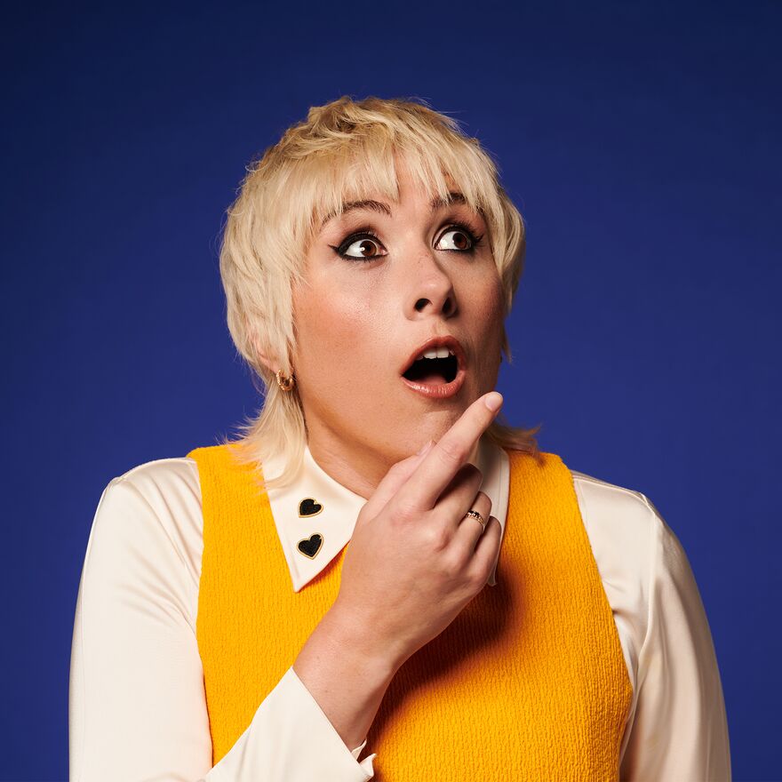 Maisie adam wearing a yellow vest and white shirt with a shocked facial expression and her hand on her chin
