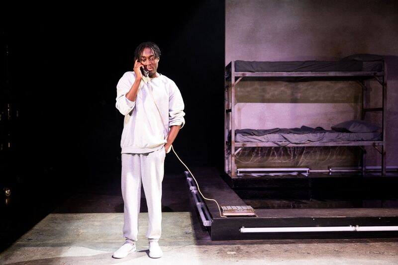 Selorm Adonu stands in the middle of the stage in a grey tracksuit, he is holding a phone up to his ear, he is in what looks like a prison cell with bunk beds in the background