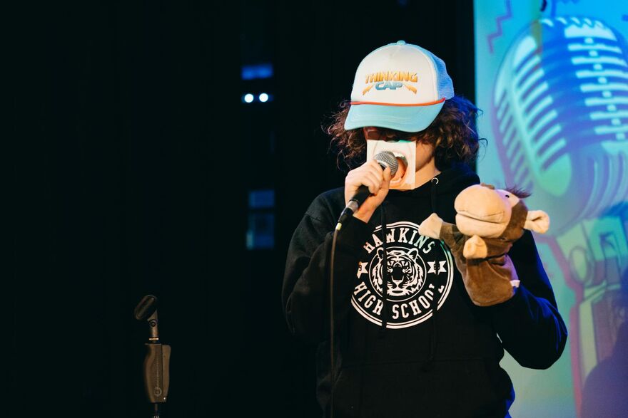A child stands on stage speaking into a microphone.They're wearing a baseball cap that says 'Thinking Cap', holding a monkey hand-puppet and have a cardboard mouth over the lower half of their face