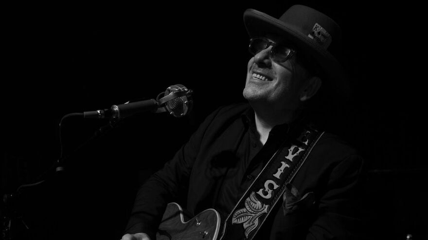 Black and White photograph of Elvis Costello Looking Up and Smiling, wearing a wide brimmed hat and holding a guitar